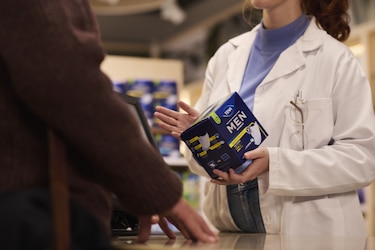 A healthcare professional in a white lab coat and blue top holds and appears to be explaining a TENA men's incontinence product package to someone partially visible in the foreground.