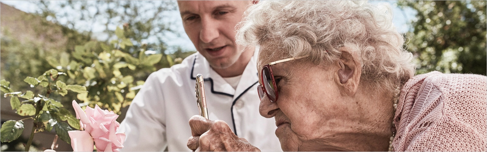 A nurse helps an elderly resident take a picture of a rose on her phone. 