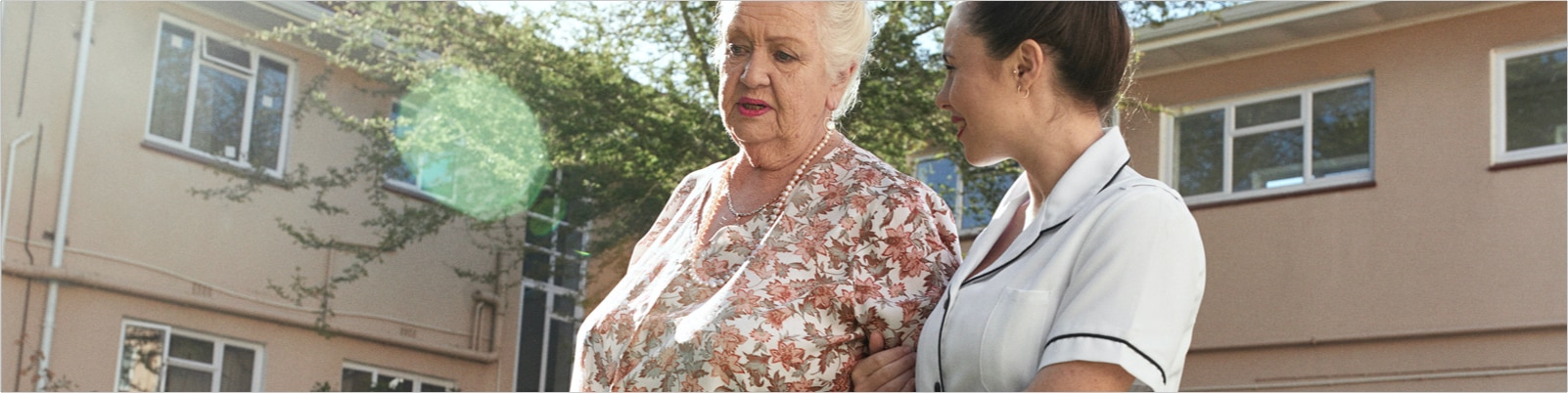 An elderly resident and her caregiver walk and talk outside a care home. 