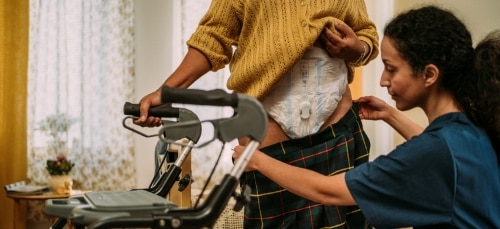 En person iklädd gul stickad tröja och rutig kjol står vid en rollator. En vårdgivare sitter på huk bredvid och hjälper till att dra upp kjolen över en TENA inkontinensprodukt.