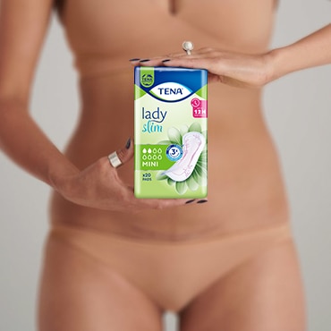 Fehér bőrű, bézs színű alsóneműt és gyűrűket viselő nő egy csomag TENA Lady Slim Mini betétet tart maga elé.