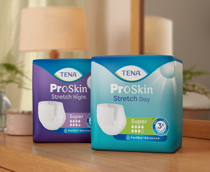 Twee verpakkingen met TENA ProSkin incontinentieproducten op een houten oppervlak. De verpakking aan de linkerkant heeft in het paars het label 'ProSkin Stretch Night', terwijl de verpakking aan de rechterkant in het blauw het label 'ProSkin Stretch Day' heeft. Op beide verpakkingen staat een afbeelding van een wit verband voor volwassenen en 'superabsorberend' staat aangegeven met meerdere waterdruppelsymbolen. Op de achtergrond is een zacht verlichte slaapkamer te zien met een lamp en een plant.