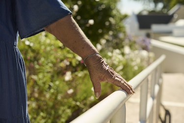 An elderly woman's hand with a bracelet rests on a white railing, with blurred garden foliage in the background.