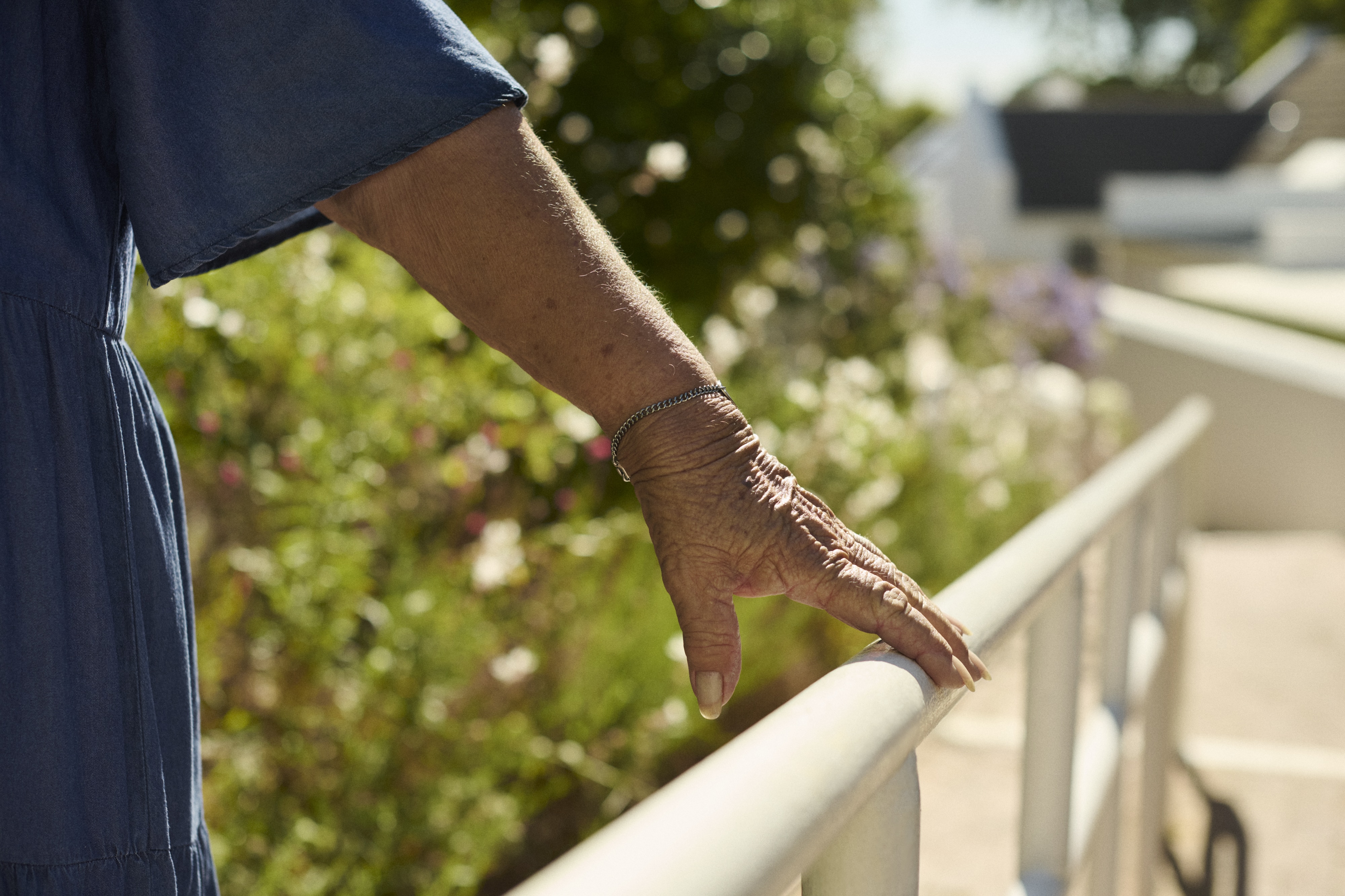 A close-up of a elderly womans hand resting on a white railing or balustrade, with blurred greenery and flowers visible in the background.