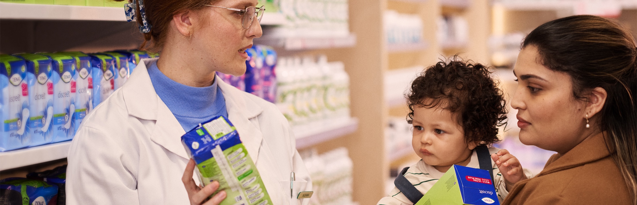 白衣の女性薬剤師が、幼い子どもを抱く母親に失禁ケアの助言をしている。二人ともTENA製品を手に持っている。