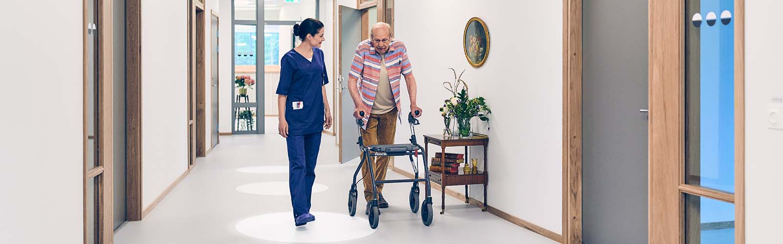 A female professional caregiver and an elderly male resident with mobility aid walking down a hallway in a nursing home environment