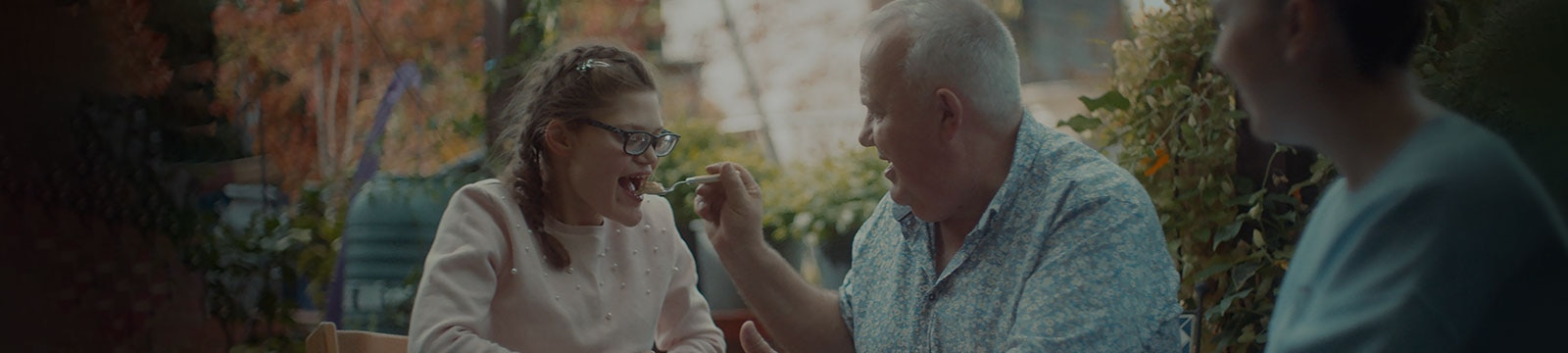 Seduto all’esterno, un uomo sorridente dà da mangiare alla figlia adolescente. 