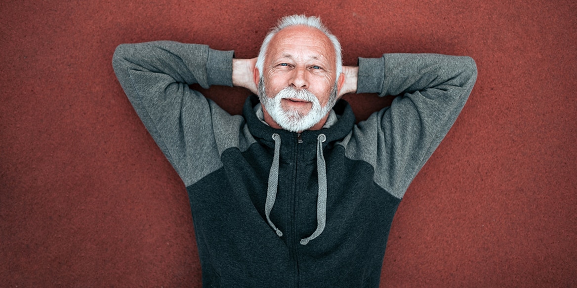 Ein Mann mit grauem Bart hat seine Hände hinter dem Kopf verschränkt und liegt auf einem roten Boden.