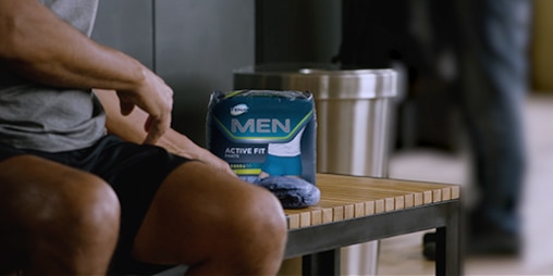 Man at the gym with a pack of TENA Men Active fit pants next to him on a bench. 