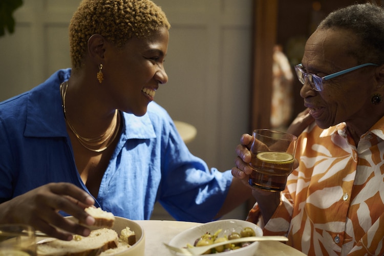 Two women share a joyful moment at a dinner table, smiling warmly at each other while enjoying food and drinks.