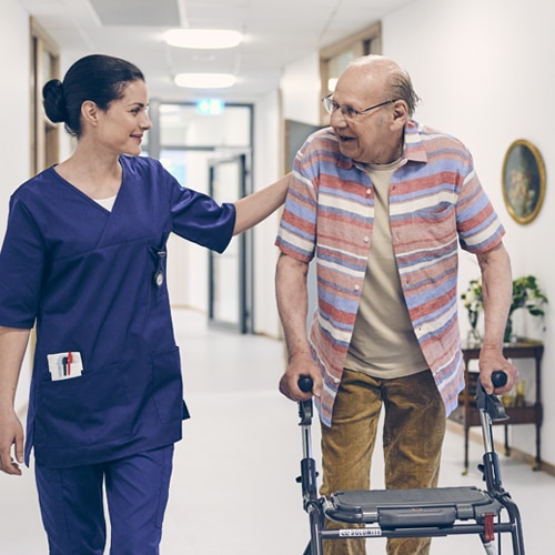 A female professional caregiver and an elderly male resident with a walking aid walk and talk in the corridor of a nursing home
