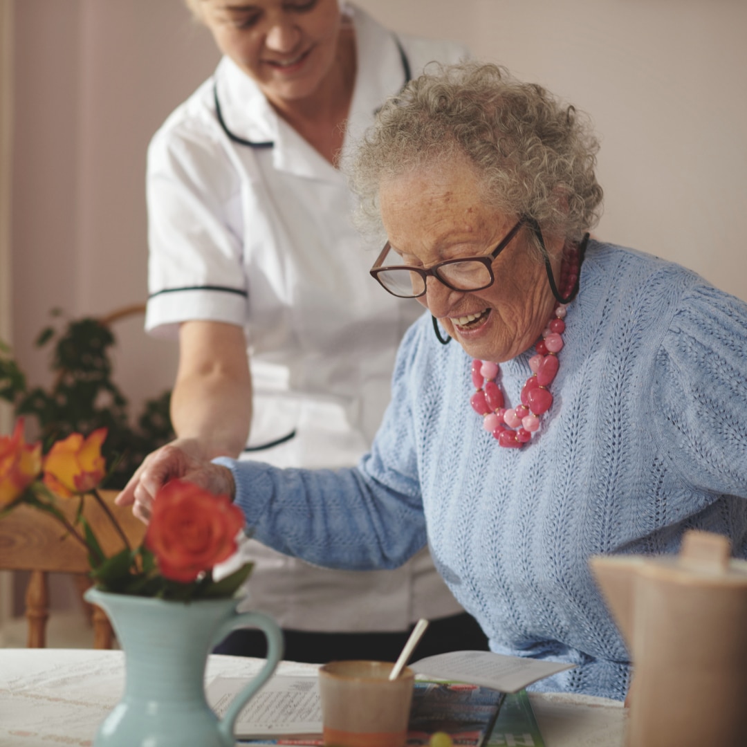A professional caregiver helps a contented nursing home resident sit down at a table with flowers in a pleasant nursing home environment.