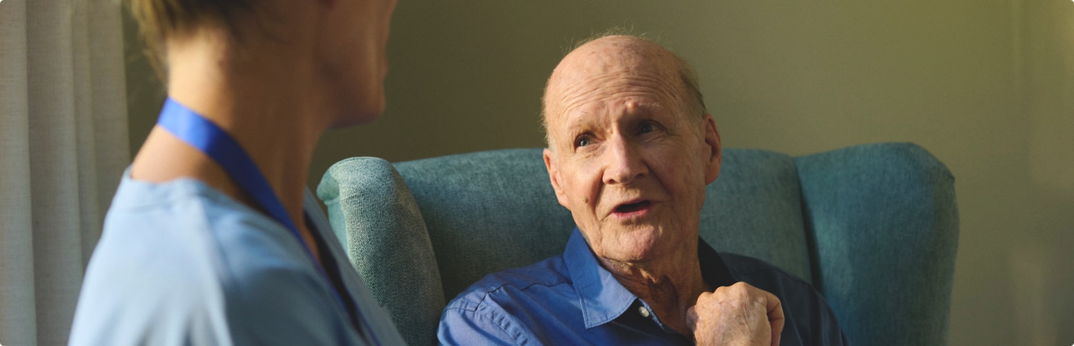 A male nursing home resident sits in a chair and has a conversation with a professional caregiver.