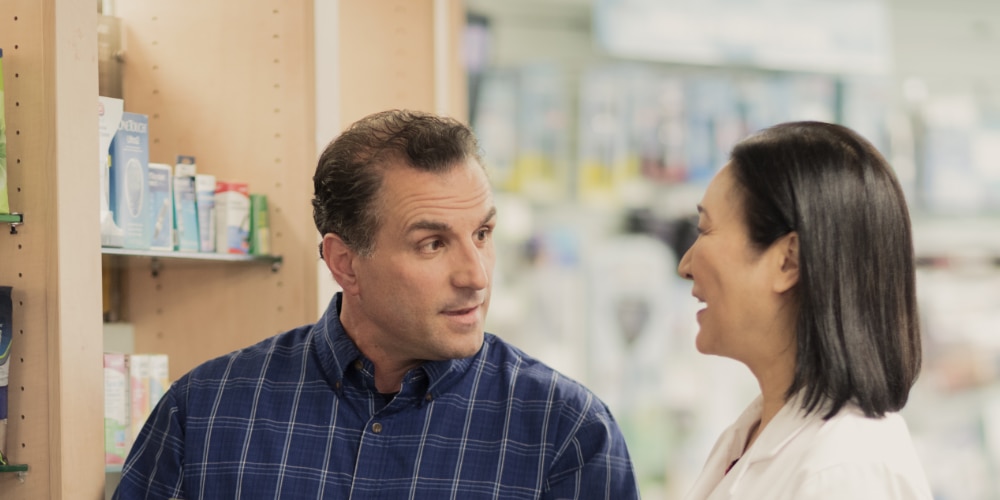 Ein Mann spricht mit einer Apothekerin im Gang einer Apotheke, TENA-Produkte sind im Regal sichtbar.