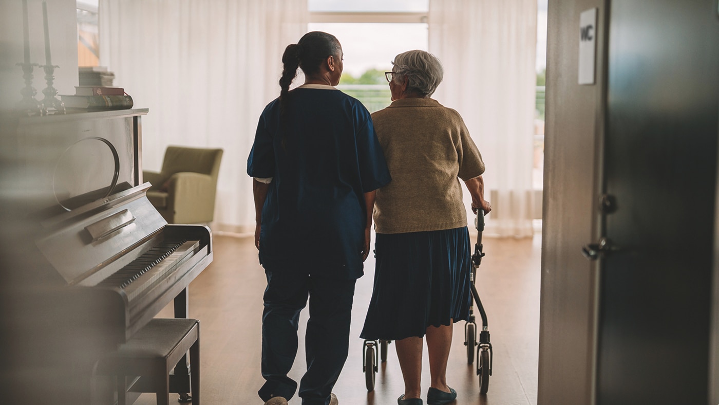 A shot taken from behind shows a professional caregiver and an elderly nursing home resident with a rollator walking down a corridor in a nursing home. 