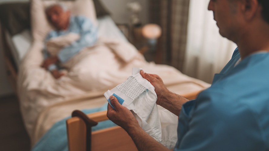 A professional caregiver holds a TENA absorbent product while a nursing home resident lies in bed. 
