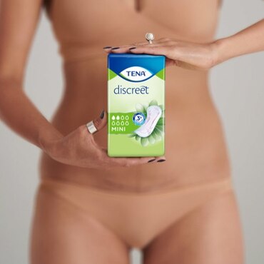 A white woman wearing beige underwear and rings, holding a TENA Discreet Mini pack in front of her.