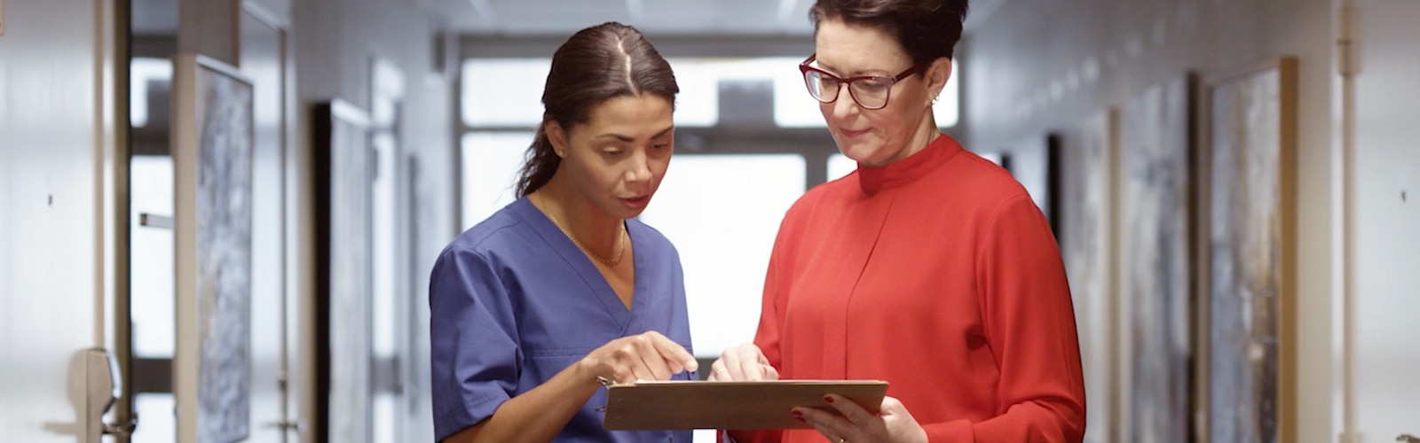 A healthcare professional in blue scrubs and a woman in a red blouse stand in a brightly lit hallway, reviewing information on a clipboard. The healthcare worker is pointing at the clipboard while the woman in red listens attentively, both focused on the discussion.