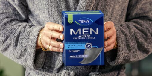 Männliches Modell im Morgenmantel, das eine Packung TENA Einlagen in der Hand hält. 