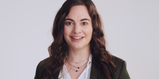 A picture of a white woman with long, brown hair, looking into the camera, smiling, with a gray background