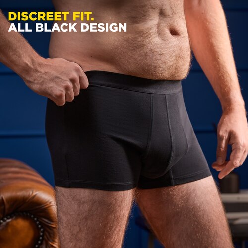 A standing man wearing only the TENA Men washable underwear and pulling them on the side at the waist. The caption at the left top corner says "Discreet fit. All Black Design".