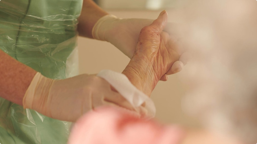 A close-up showing a professional caregiver as they gently clean the skin on the lower arm of an elderly nursing home resident. The caregiver is wearing protective gloves and protective apron.