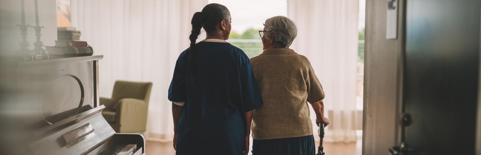 A female professional caregiver reassuringly supports a female nursing home resident as they walk and talk in a nursing home. There is a piano nearby and the resident has a rollator.
