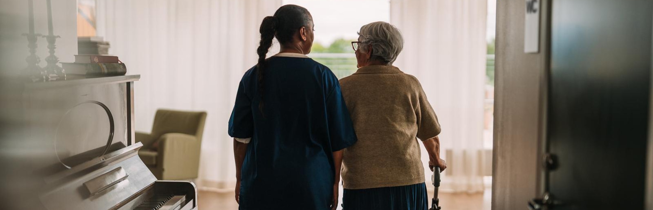 A female professional caregiver reassuringly supports a female nursing home resident as they walk and talk in a nursing home. There is a piano nearby and the resident has a rollator.