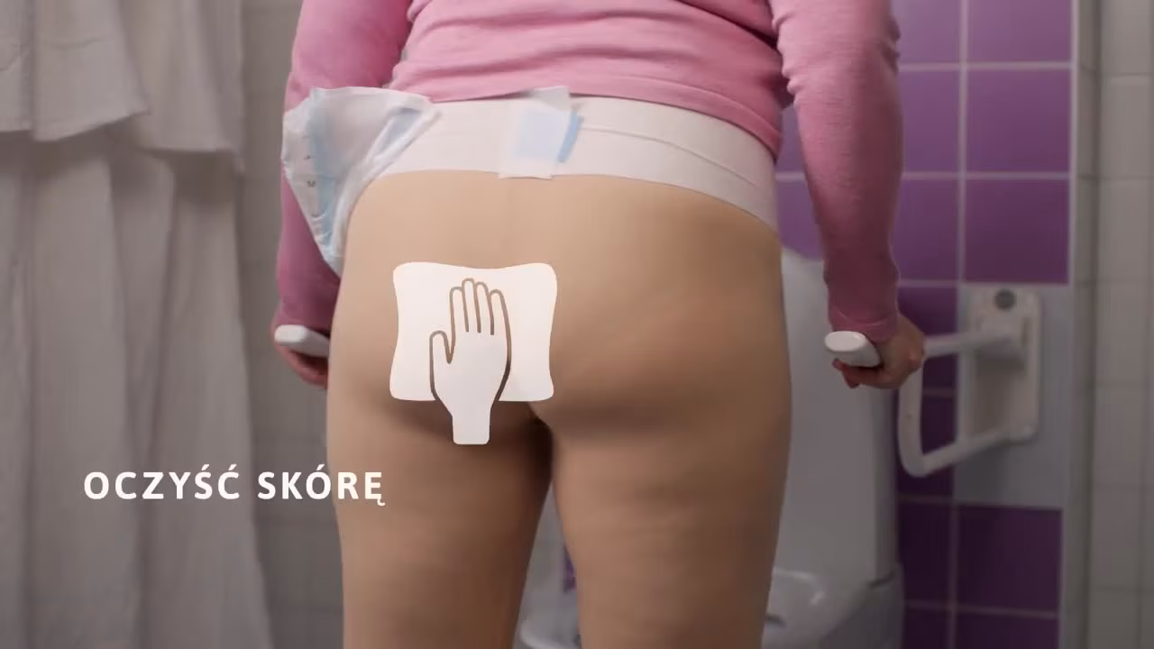 Zbliżenie osoby siedzącej na toalecie z uchwytami wspomagającymi, ubraną w jednorazowe pieluchomajtki dla dorosłych i pończochy uciskowe. Ich dłonie trzymają się uchwytów dla podparcia. W tle widoczne są białe ściany wyłożone kafelkami, co wskazuje na łazienkowe otoczenie.