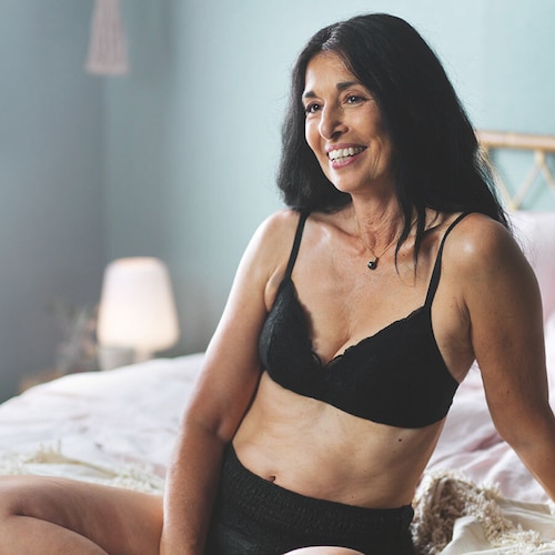 Vrouw met lang donker haar zit lachend op bed en draagt zwart TENA Silhouette-ondergoed, ontworpen voor comfort en bescherming.