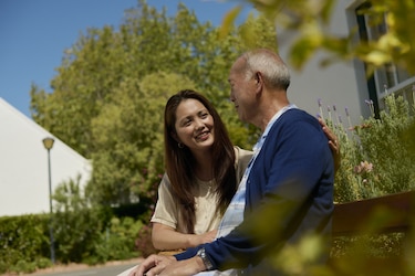 A young woman with long brown hair and an elderly man with short gray hair sit together on a wooden bench in a lush garden. The woman is smiling warmly while placing her hand on the man's shoulder, and they are engaged in a pleasant conversation.