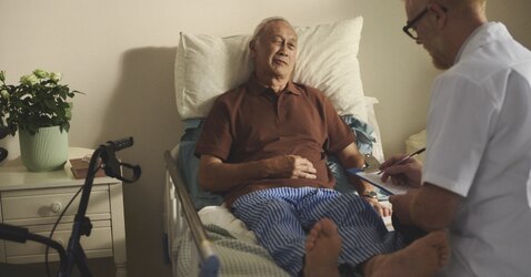 An elderly male resident laid in bed with a healthcare professional at the end of his bed making notes