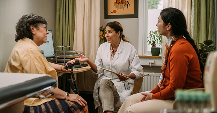 In a nursing home room with green curtains and indoor plants, a healthcare professional in white scrubs sits between two women, gesturing while engaged in conversation. The room is warmly lit and decorated with framed artwork on the wall.
