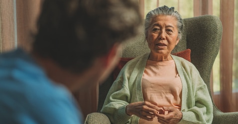 An elderly woman wearing a pale green cardigan over a peach-colored top sits in a green armchair, engaged in conversation with someone visible from behind in the foreground. She appears to be speaking and gesturing with her hands.