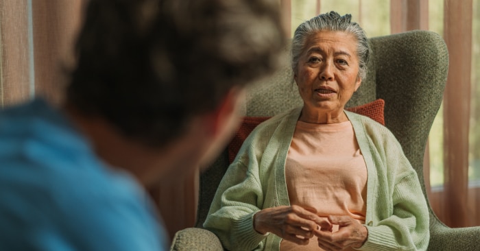 An elderly woman wearing a pale green cardigan over a peach-colored top sits in a green armchair, engaged in conversation with someone visible from behind in the foreground. She appears to be speaking and gesturing with her hands.