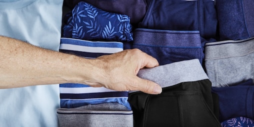 Eine männliche Hand wählt ein Paar waschbare TENA Boxershorts aus einem Stapel normaler Herrenunterwäsche. 