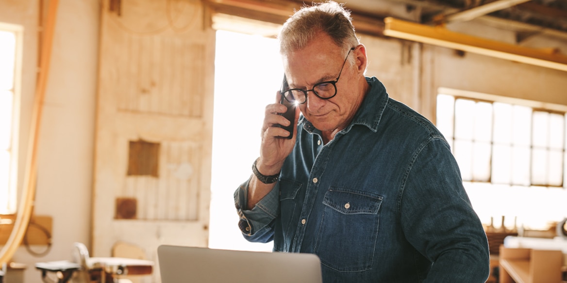 Ein Mann mit Brille steht und schaut in sein Notebook während er ein Telefon an sein rechtes Ohr hält.