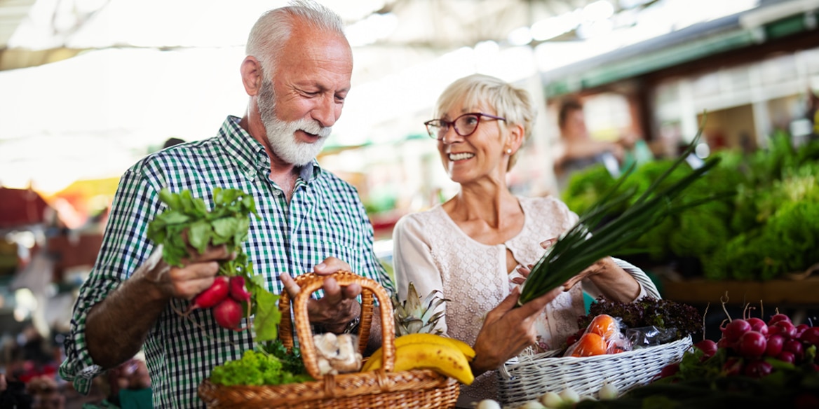 Ein Seniorenpaar beim Markt-Einkauf hält Körbe mit Obst und Gemüse in den Händen.