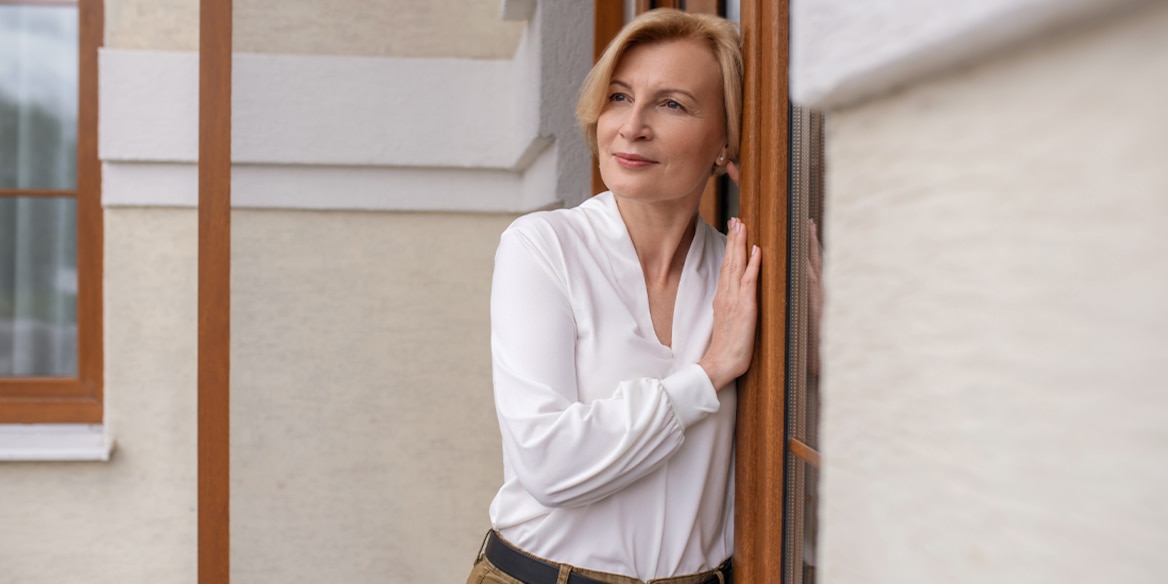 Eine blonde Frau in weißer Bluse lehnt gedankenverloren am Türrahmen