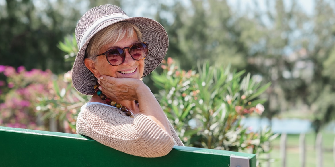 Eine Frau mit Sonnenbrille und Hut sitzt bei Sonnenschein im Garten auf einer grünen Bank und lächelt