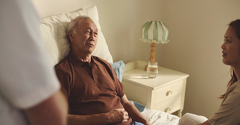 An elderly man in a bed inside talking to a female caregiver. 