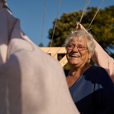 Mujer mayor sonriente en un espacio exterior rodeada de ropa colgada en un tendal.