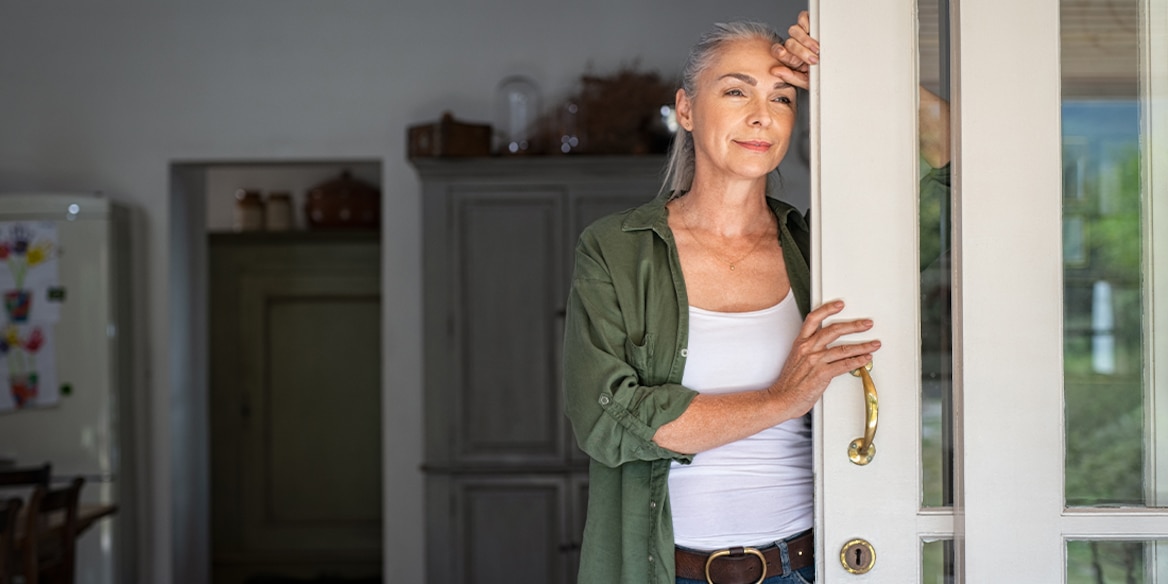 Eine Frau mit grauen Haaren steht nachdenklich an einer Tür und hält sich fest.