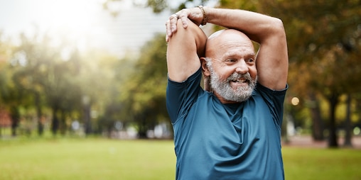 Ein fröhlicher Mann mit grauem Bart in Sportkleidung steht im Park und dehnt seine Arme über dem Kopf.