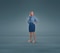 A woman standing against a plain blue background with both hands on her hips, dressed in a blue shirt, blue skirt, and orange shoes, wearing Knee-high JOBST Relief compression stockings.