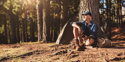Ein Mann mit Basecap, kurzen Hosen und Wanderschuhen lehnt im Wald an einem Baum vor einem Zelt