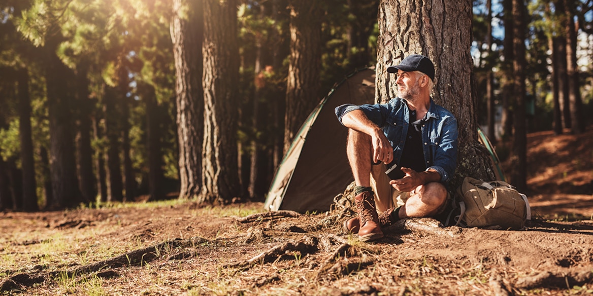 Ein Mann mit Basecap, kurzen Hosen und Wanderschuhen lehnt im Wald an einem Baum vor einem Zelt.