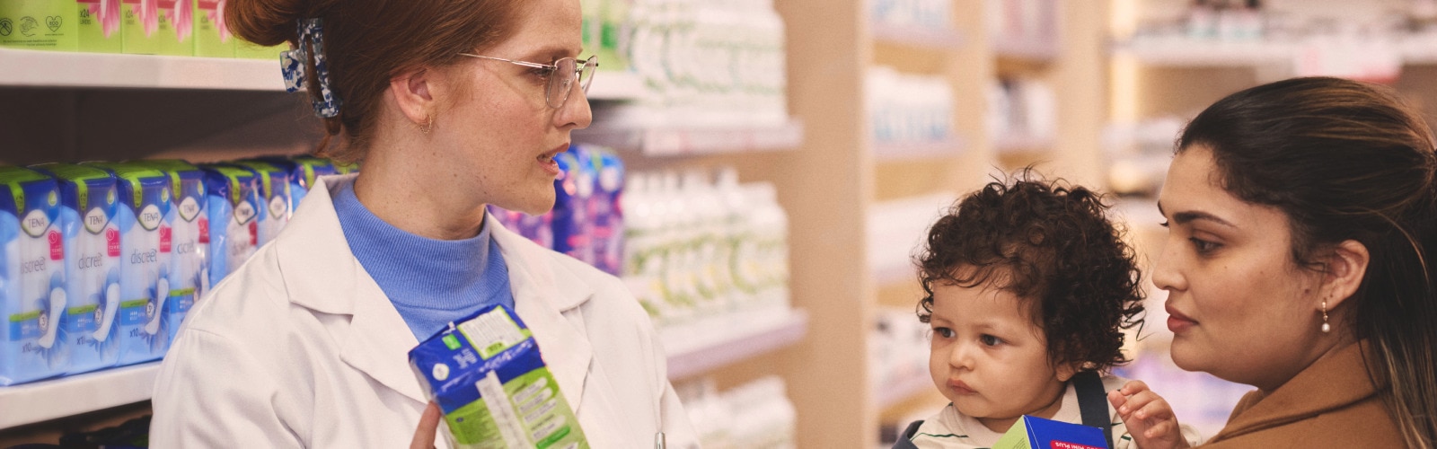 A female pharmacist recommends a TENA incontinence product to a mother who is carrying a young child 