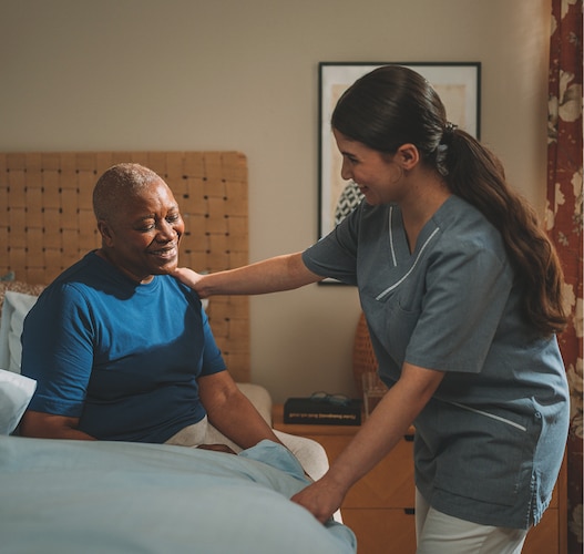 A female professional caregiver talks and smiles with an elderly female nursing home resident in the resident’s room