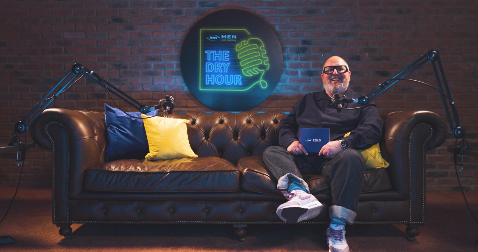 Comedian Justin Moorhouse sat on a sofa in front of Dry Hour TENA logo